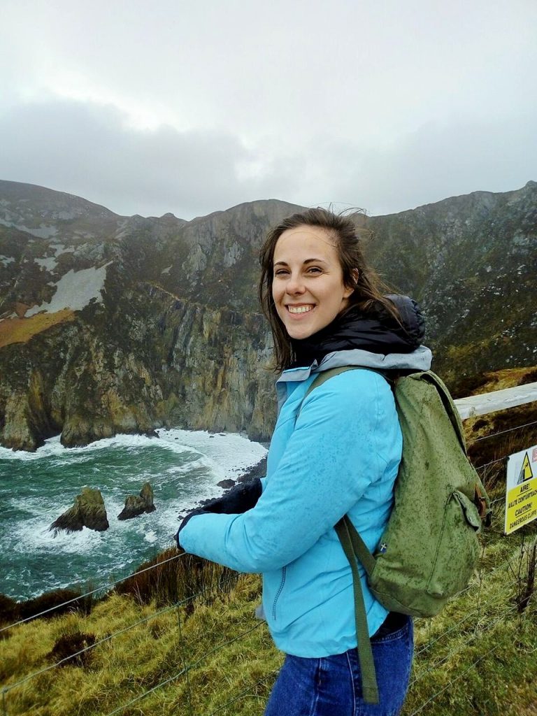 Smiling woman in a blue jacket with a green backpack standing by a rocky coastline and cliff railing.