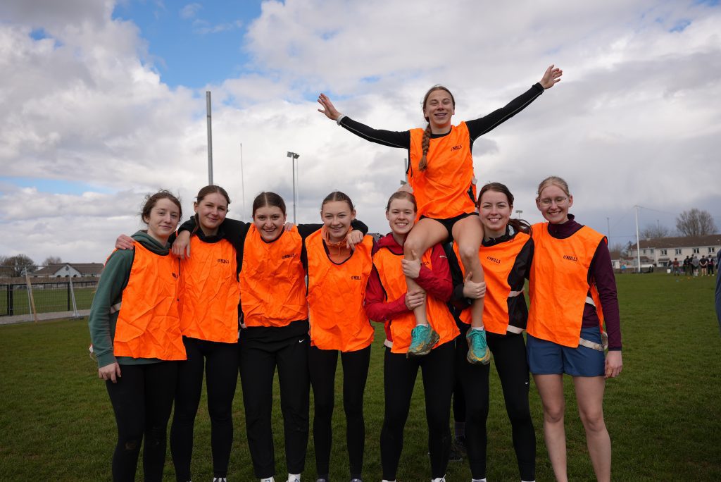 A group of smiling young athletes in bright orange vests posing together on a grass field, one girl being lifted on teammates' shoulders with arms raised.