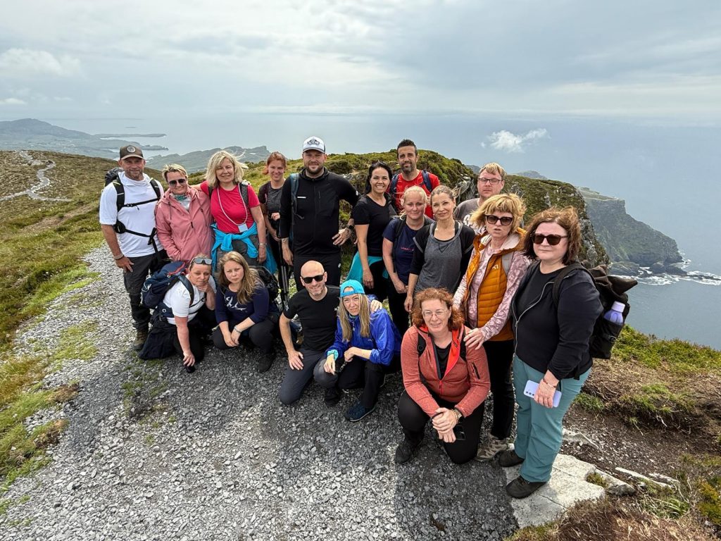 Group of about 16 hikers posing on a rocky coastal trail with the sea and cliffs in the background, under a cloudy sky.