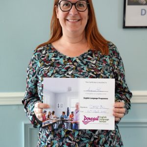 Smiling woman in a floral top holds a certificate for an English language programme indoors.