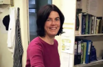 Smiling woman in a pink sweater stands in a cluttered office with shelves of binders in the background.