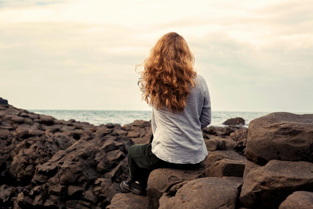 Person with long curly hair sits on dark rocks by the ocean, facing away from the camera.