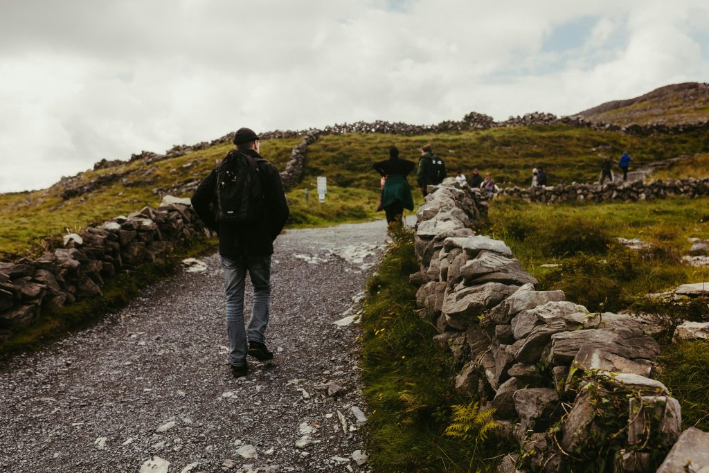 Man with a backpack walking away on a rocky gravel path flanked by stone walls in a hilly, green countryside under a cloudy sky.