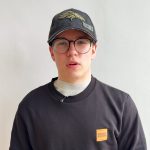 Young person wearing a dark baseball cap, black glasses, and a black sweatshirt with an orange square logo, facing the camera.