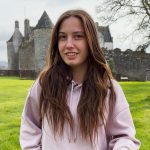 Young woman with long brown hair in a pale pink hoodie standing on a green lawn with a historic stone castle in the background.
