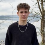 Young man in a black knit sweater and silver chain stands by a calm lake with bare trees behind him.