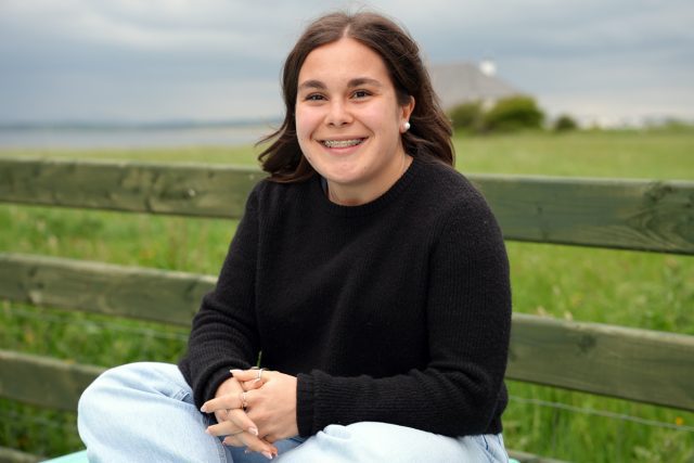 Smiling teenage girl with braces sits on a wooden bench in a grassy field by the coast, wearing a black sweater and light jeans.