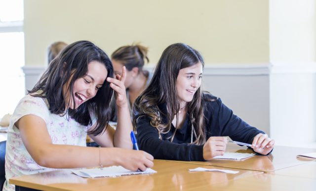 Two smiling girls sit at a desk, writing on papers in a classroom setting, collaborating on work.