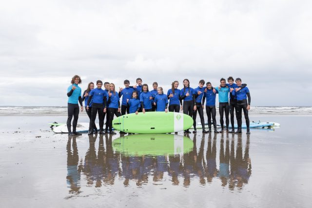 Group of surfers in blue wetsuits posing on a wet beach with green surfboards in front of them.