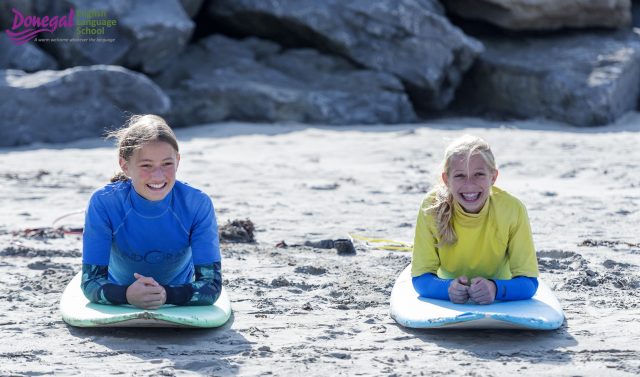 Two smiling girls sit on surfboards on a sandy beach, wearing blue and yellow rash guards, with rocky cliffs behind them.