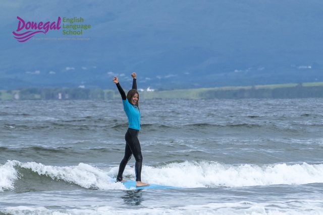 Person wearing a blue wetsuit stands on a surfboard in the sea with waves near shore; logo for 'Donegal English Language School' in the top-left corner
