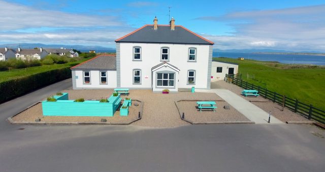 White two-story house with gray trim and turquoise outdoor furniture in a gravel yard, sea and hills in background.