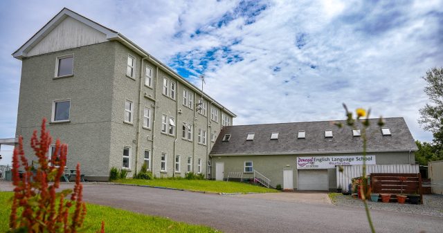 Exterior view of a pale green, multi-story school building under a cloudy blue sky; sign reads 'Donegal English Language School' on the lower right.