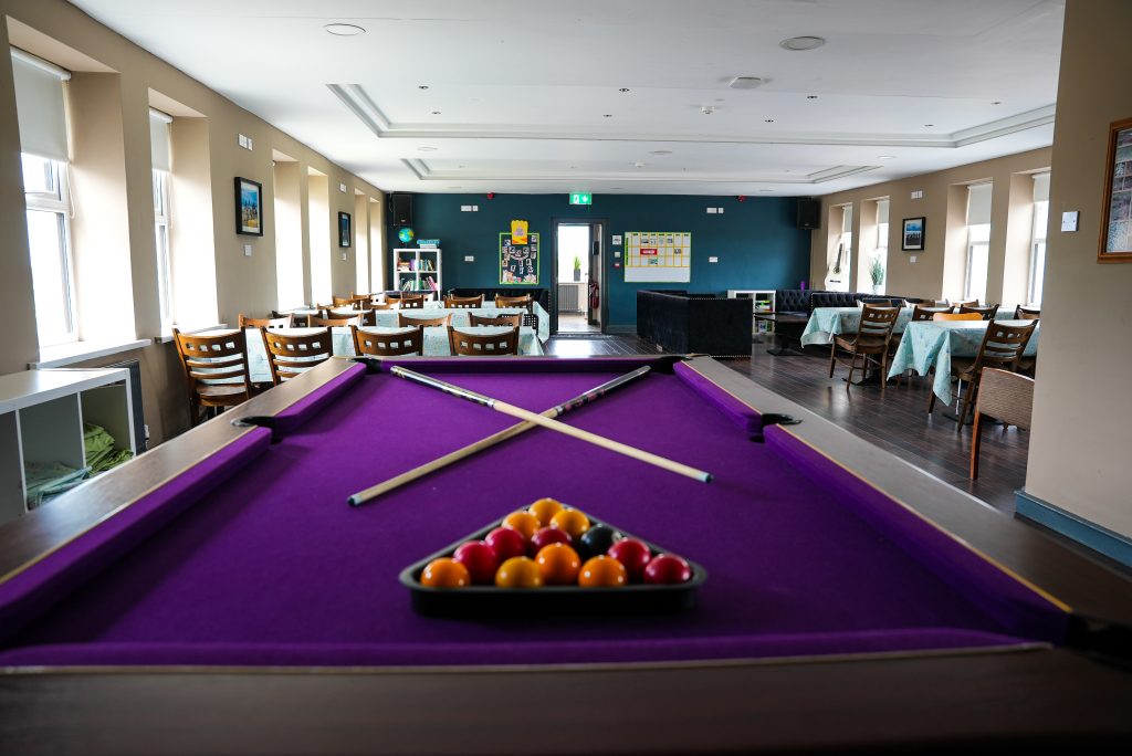 Pool table with purple felt in a bright lounge; cue sticks rest on the table with a rack of balls at the edge.