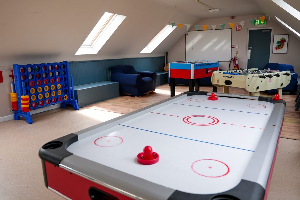 Recreation room with an air hockey table in the foreground, plus a foosball table and shuffleboard nearby, and blue seating along the wall.