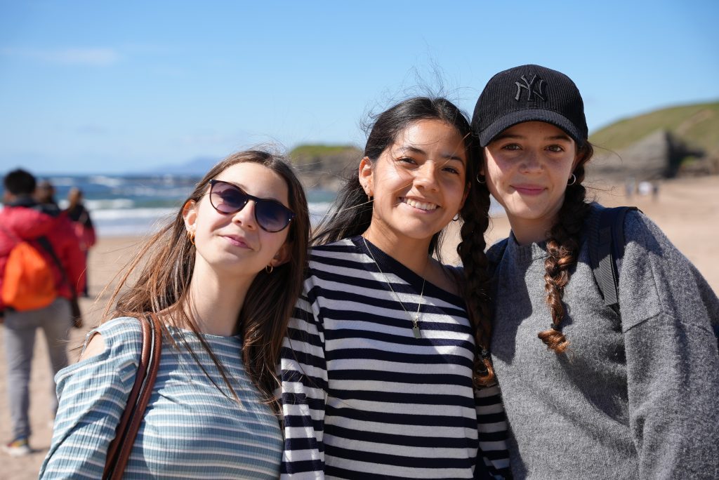 Three friends at the beach posing for a selfie, smiling with the ocean and blue sky behind them.