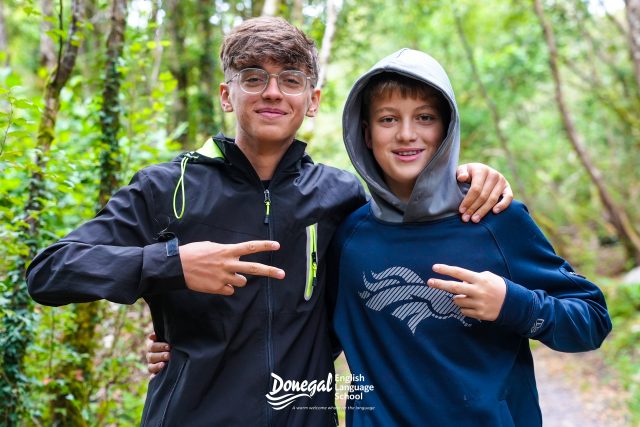 Two boys with arms around each other, smiling in a forest path.