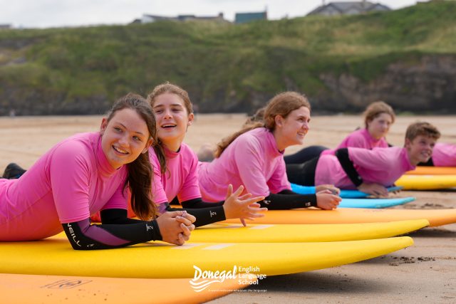 Group of smiling people in pink shirts lying on colorful surfboards on a sandy beach, with cliffs in the background and a visible Donegal English Language School logo on the board.